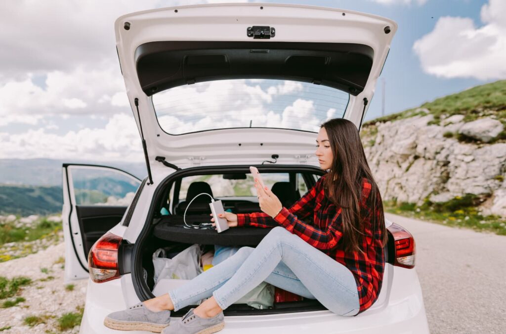 Mujer sentada en el baúl abierto de un auto, con un celular en la mano