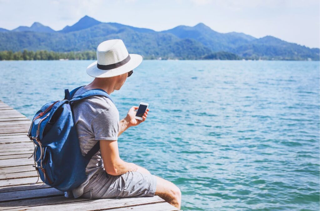 Hombre con sombrero sosteniendo un celular en la mano, sentado en un muelle frente a un lago